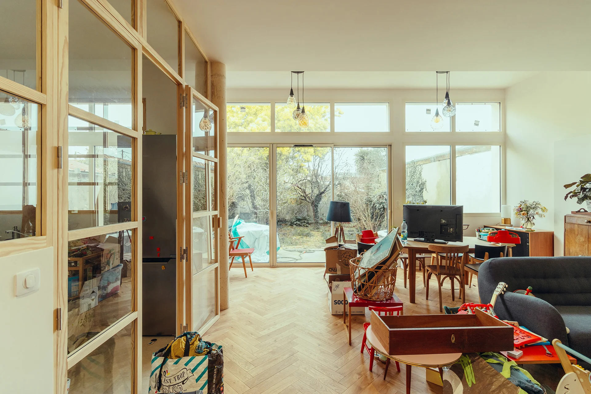 Vue du salon en double hauteur, grande lumière et cuisine séparée par une verrière en pin
