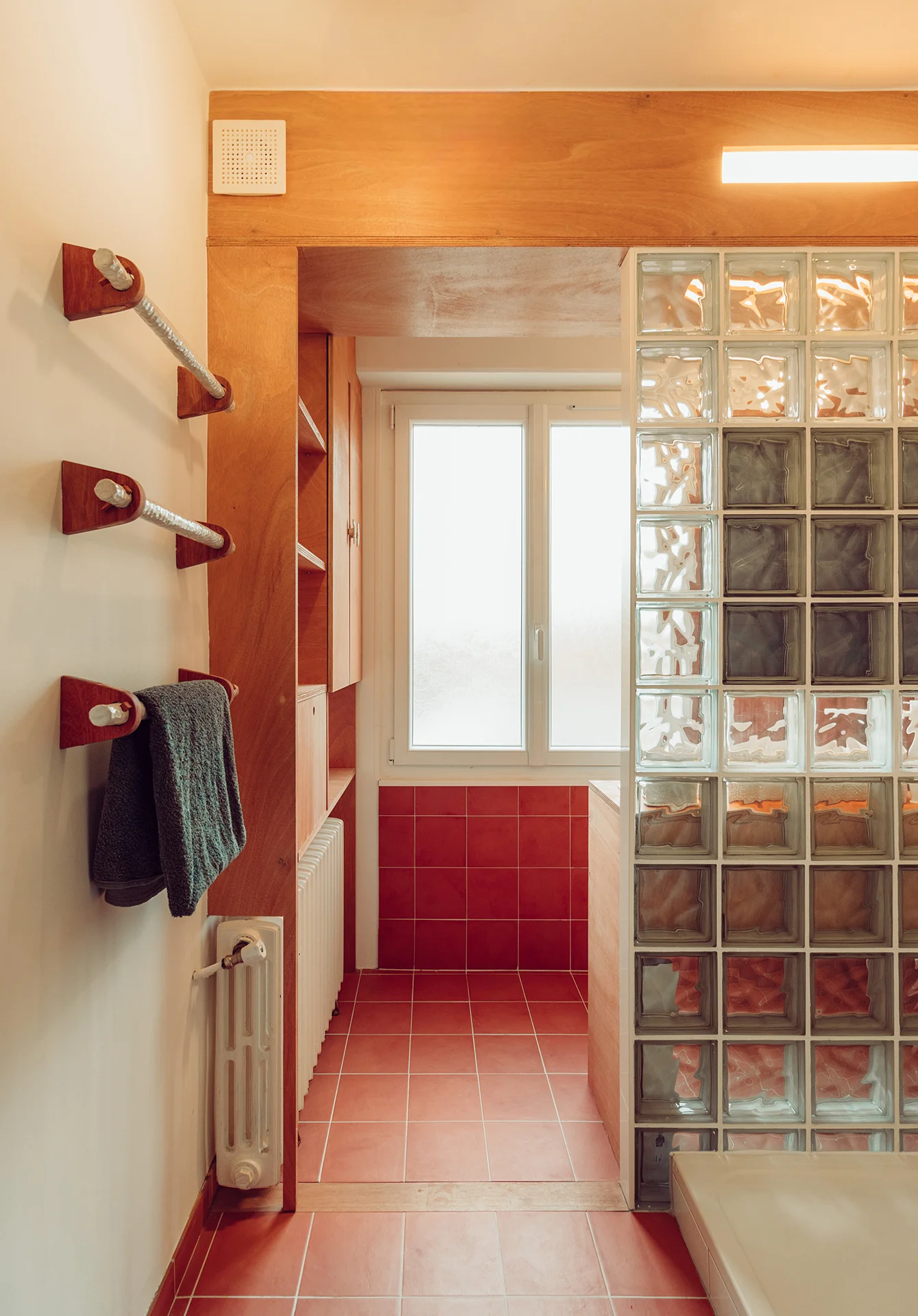 Vue de la salle de bain depuis l'entrée, deux parties séparées par la cloison en pavés de verre qui s'articule avec un caisson en bois de distribution de l'éclairage et de l'aération. Au premier plan, accroches serviettes sur mesure du céramiste Hugo Gomez (DTR)