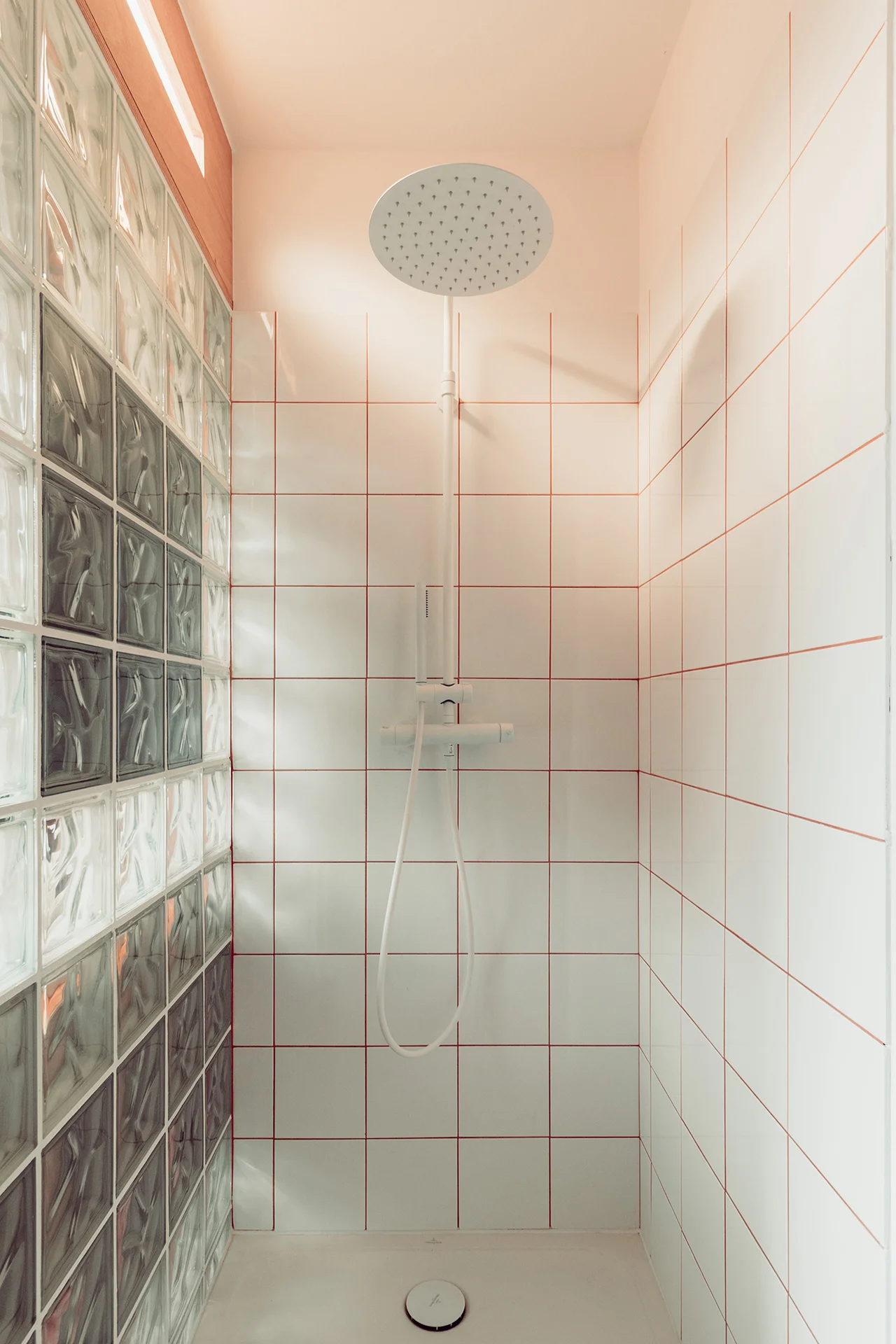 Cabine de douche en négatif du reste (carrelage blanc joints terracotta), colonne de douche blanc mat, éclairage intégré à la douche depuis le caisson en bois